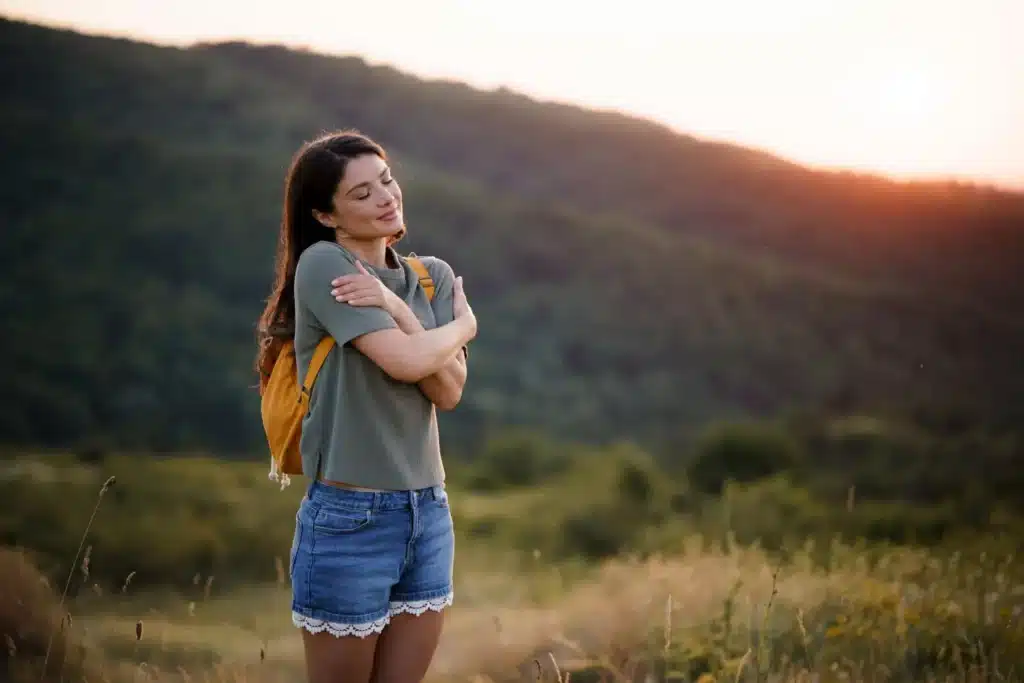 Femme s'enlaçant elle-même dans un paysage naturel, symbole d'accueil de soi, de douceur et de réconciliation intérieure.